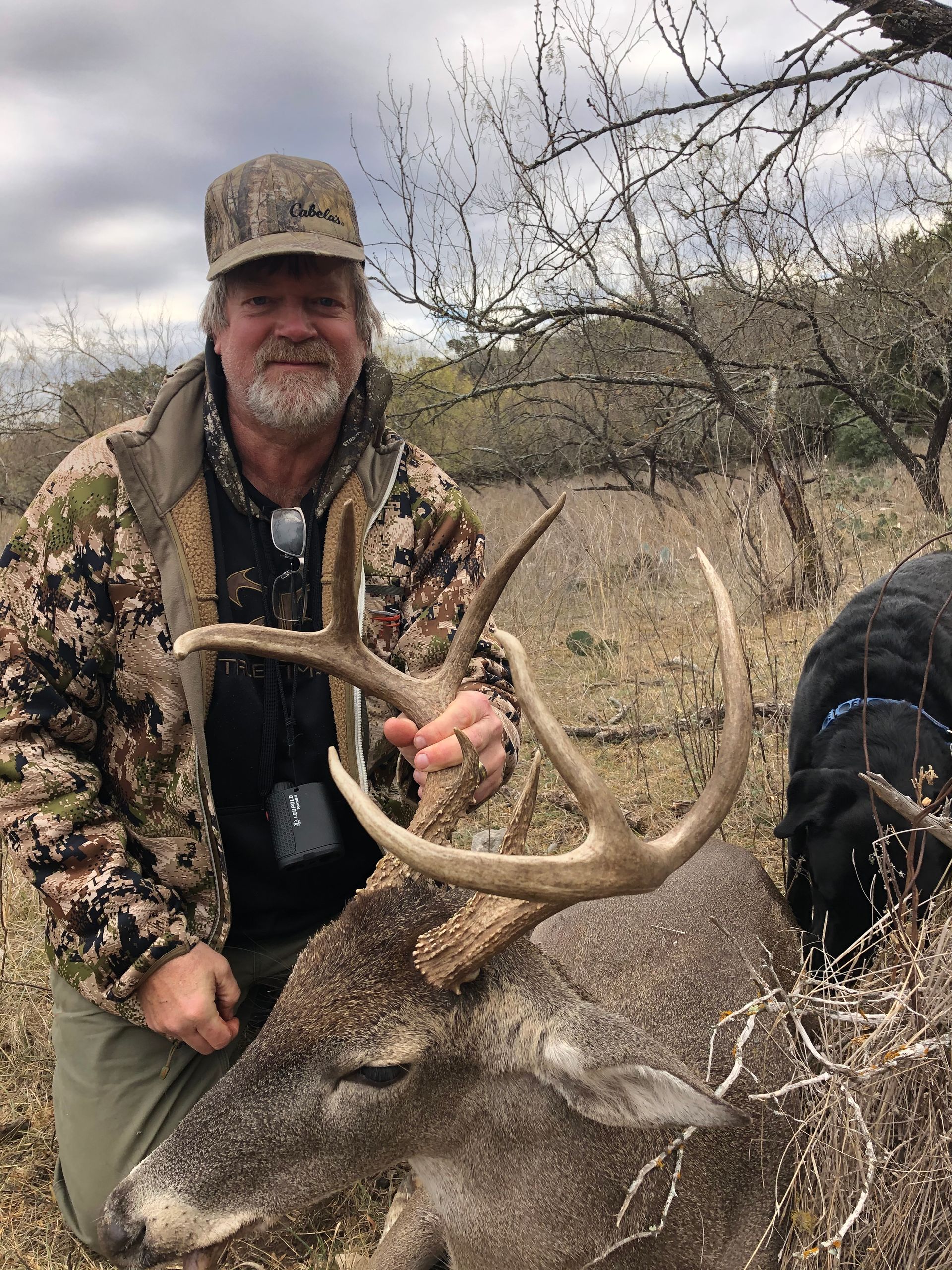 Man in camouflage holding large buck's antlers, dog nearby, outdoor setting with bare trees.