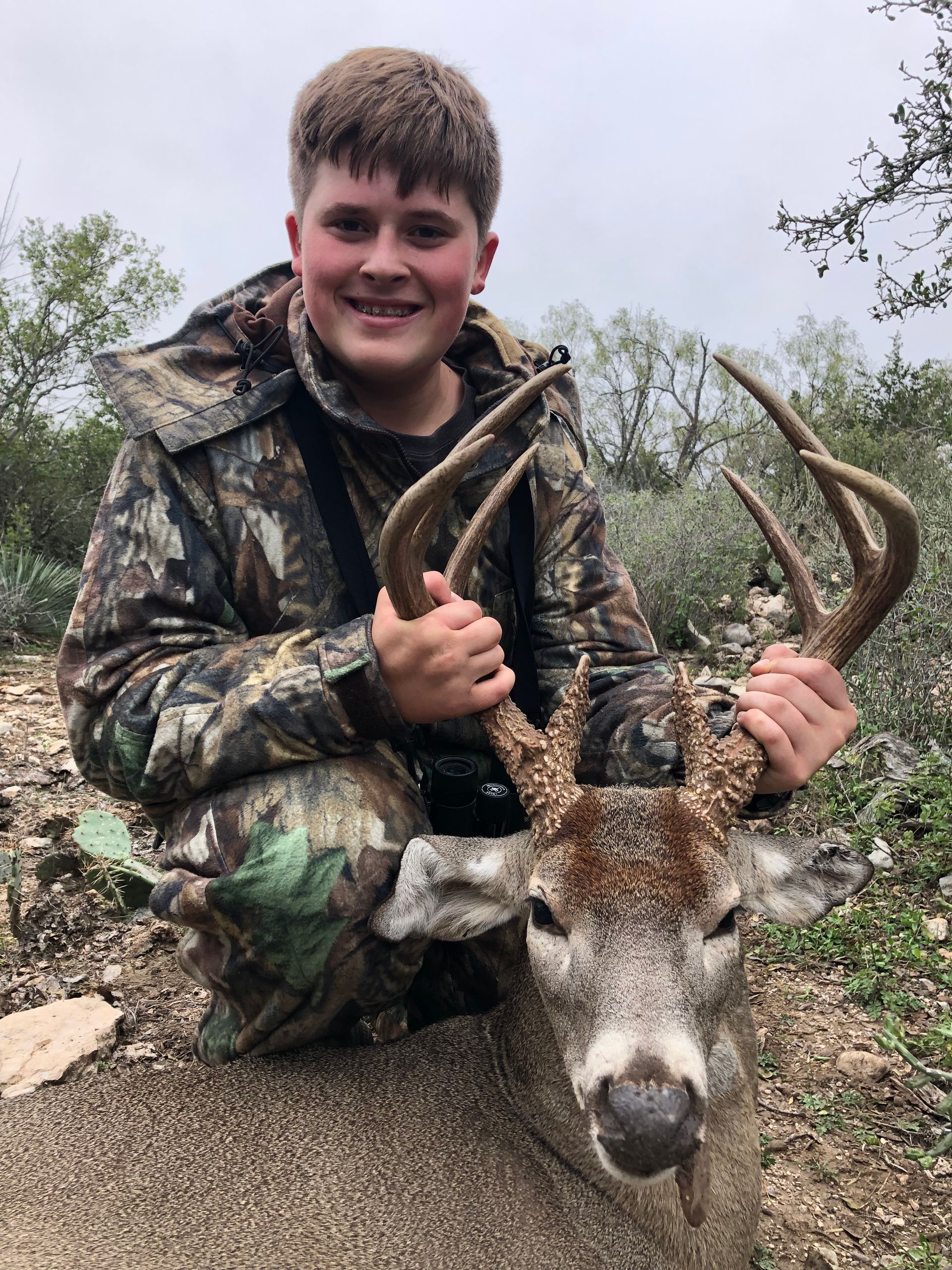 Boy in camouflage kneels, smiling, holding a deer's large antlers. Outdoor setting, cloudy sky.