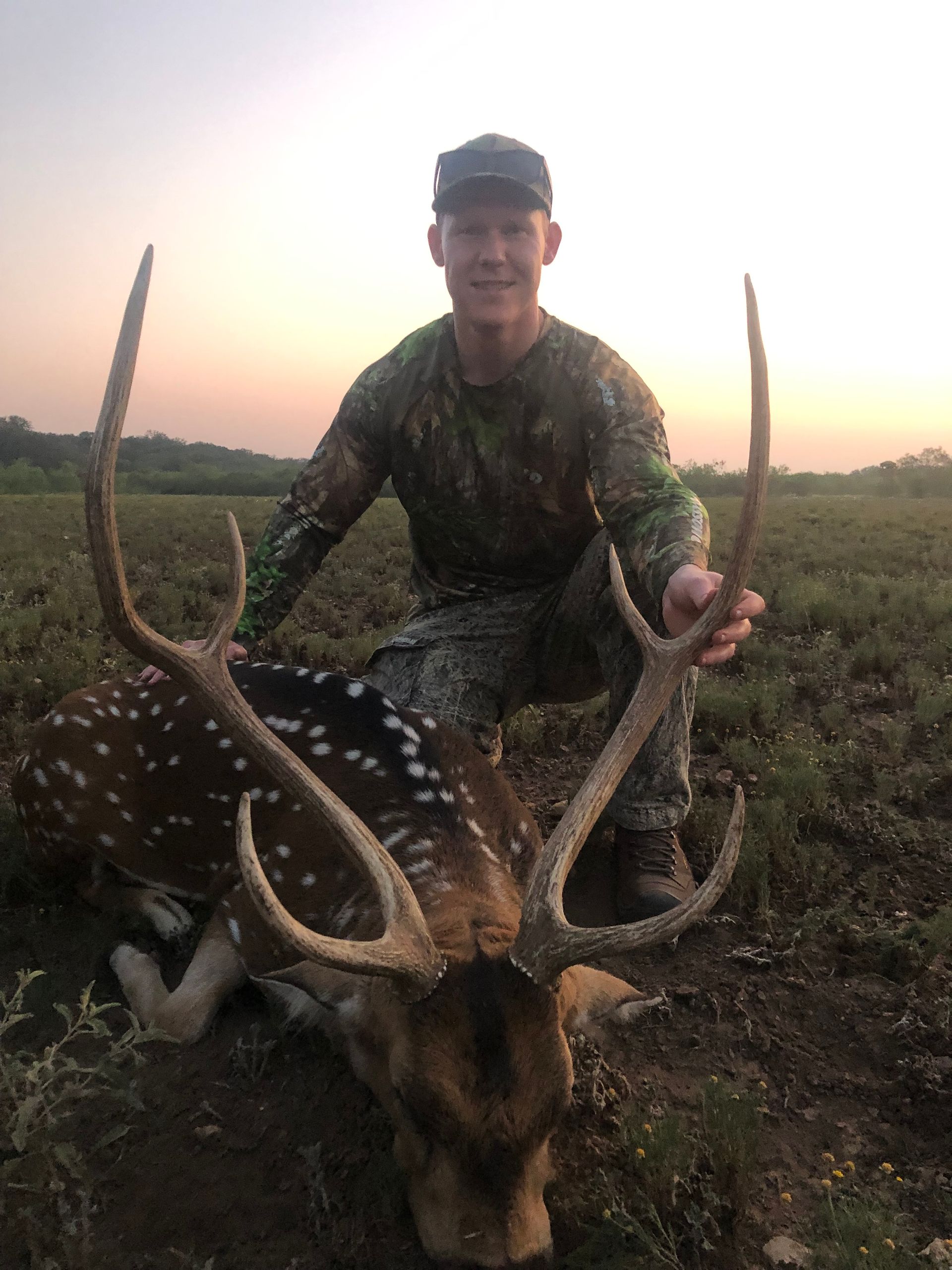 Hunter kneeling beside a spotted deer with large antlers in a field at sunset.