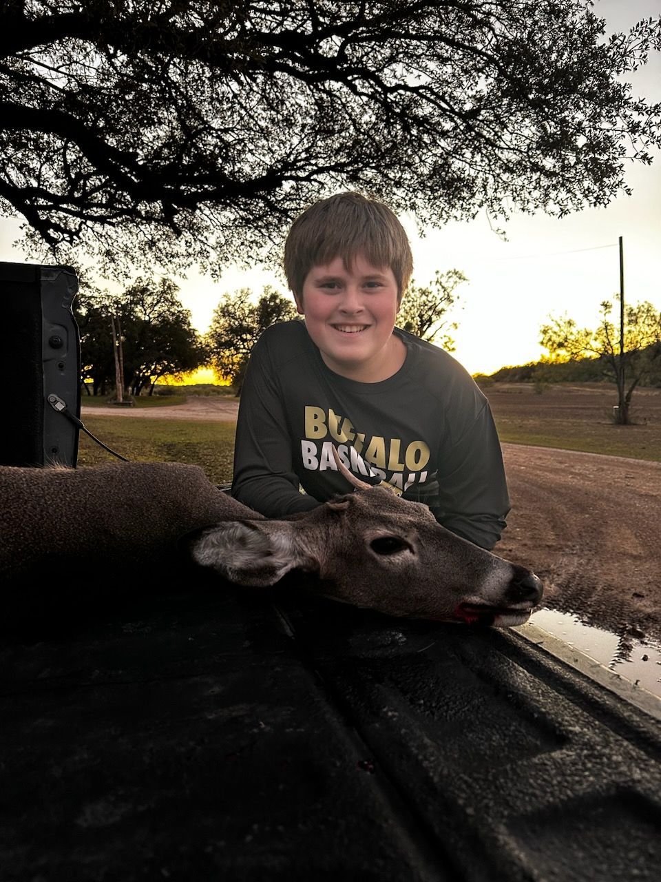 Boy smiles next to a deer on a truck bed at dusk, under a tree.