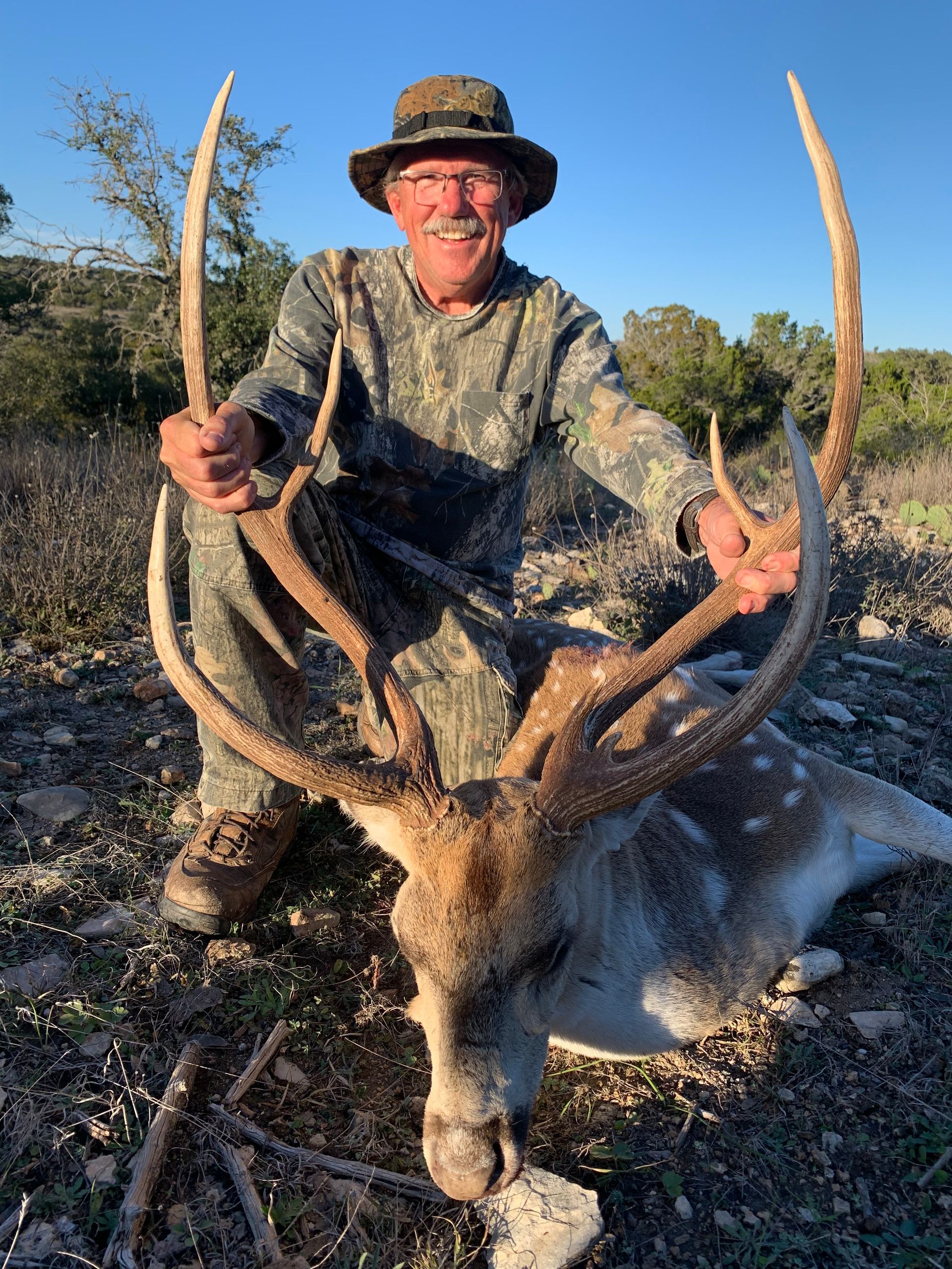 Hunter in camo with a large spotted deer. Holding antlers, smiling. Outdoors, sunny.