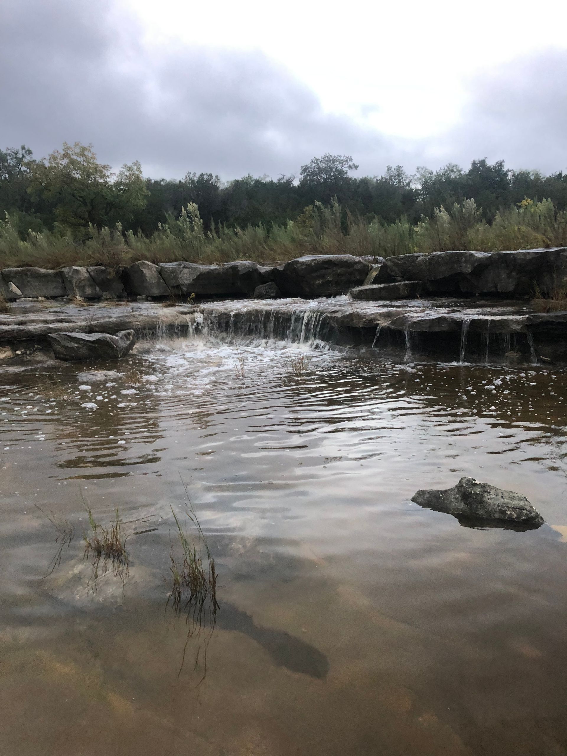 Water cascading over rocks into a calm, shallow pool. Trees line the bank under an overcast sky.