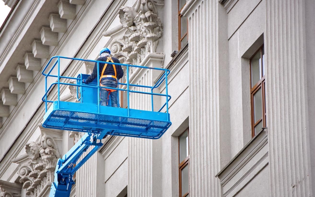 Worker in a blue lift basket repairs building facade, wearing safety harness.
