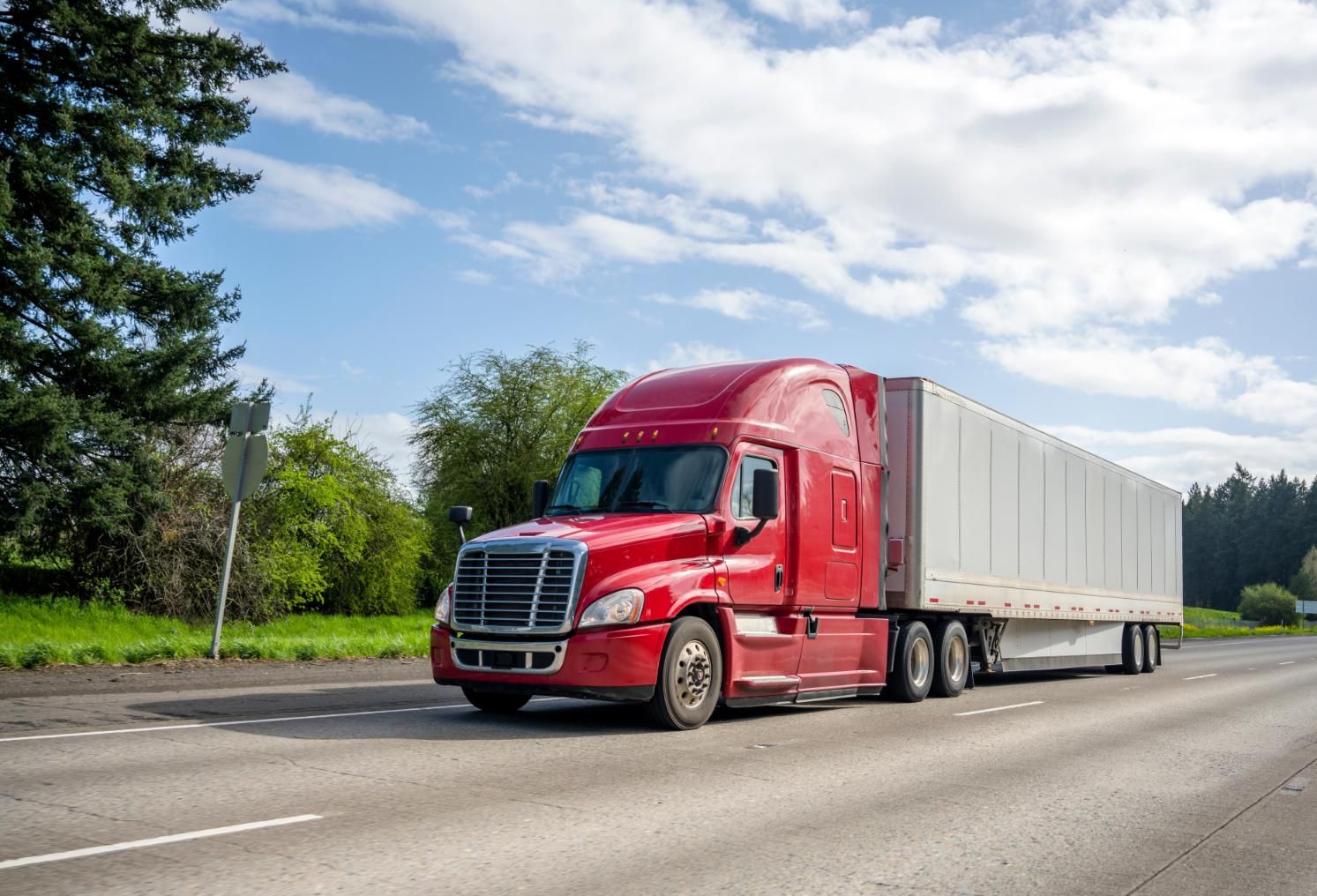 Red semi-truck driving on a road, hauling a white trailer, under a blue sky.