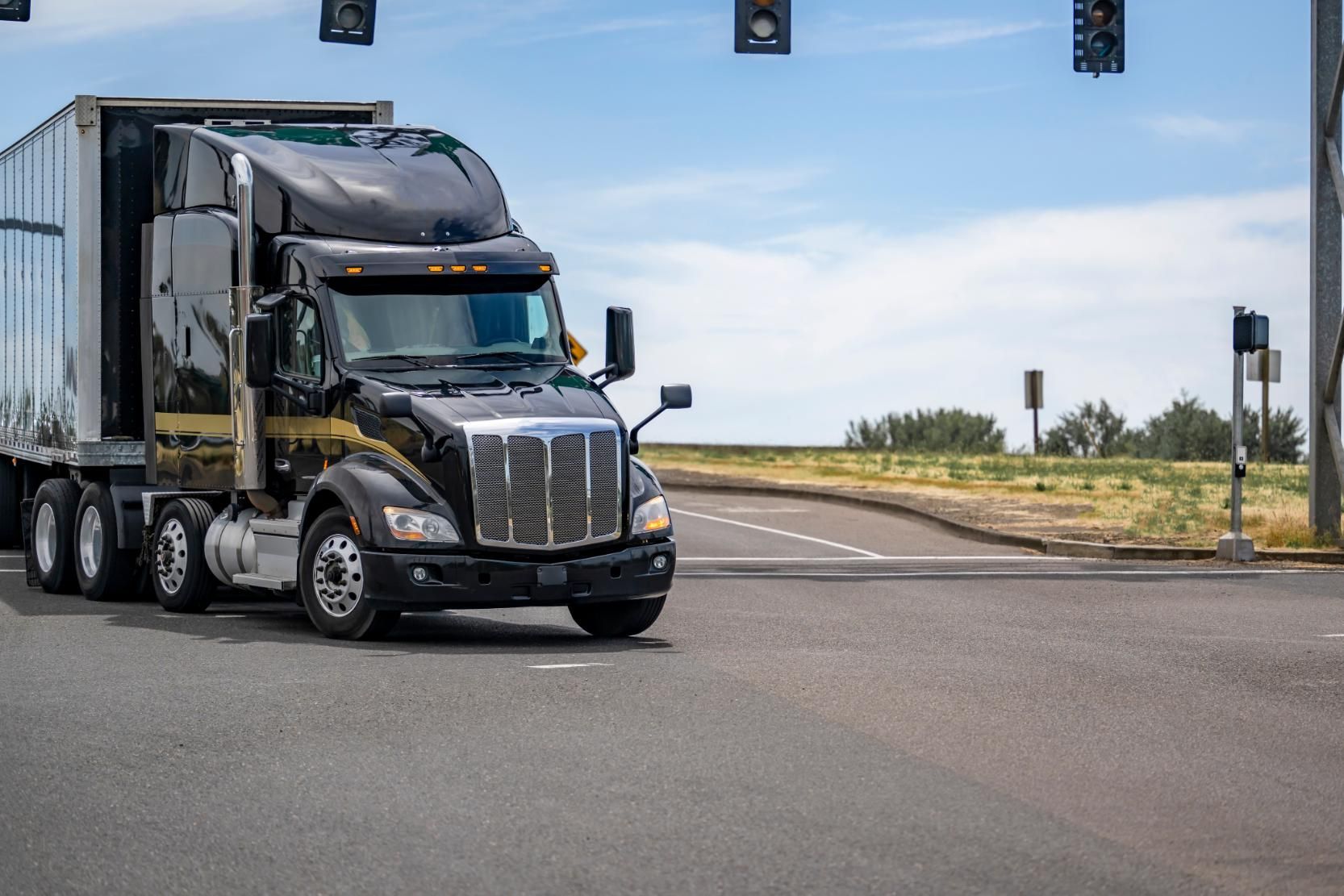 Black semi-truck with a trailer driving on a road near traffic lights and a grassy hillside.