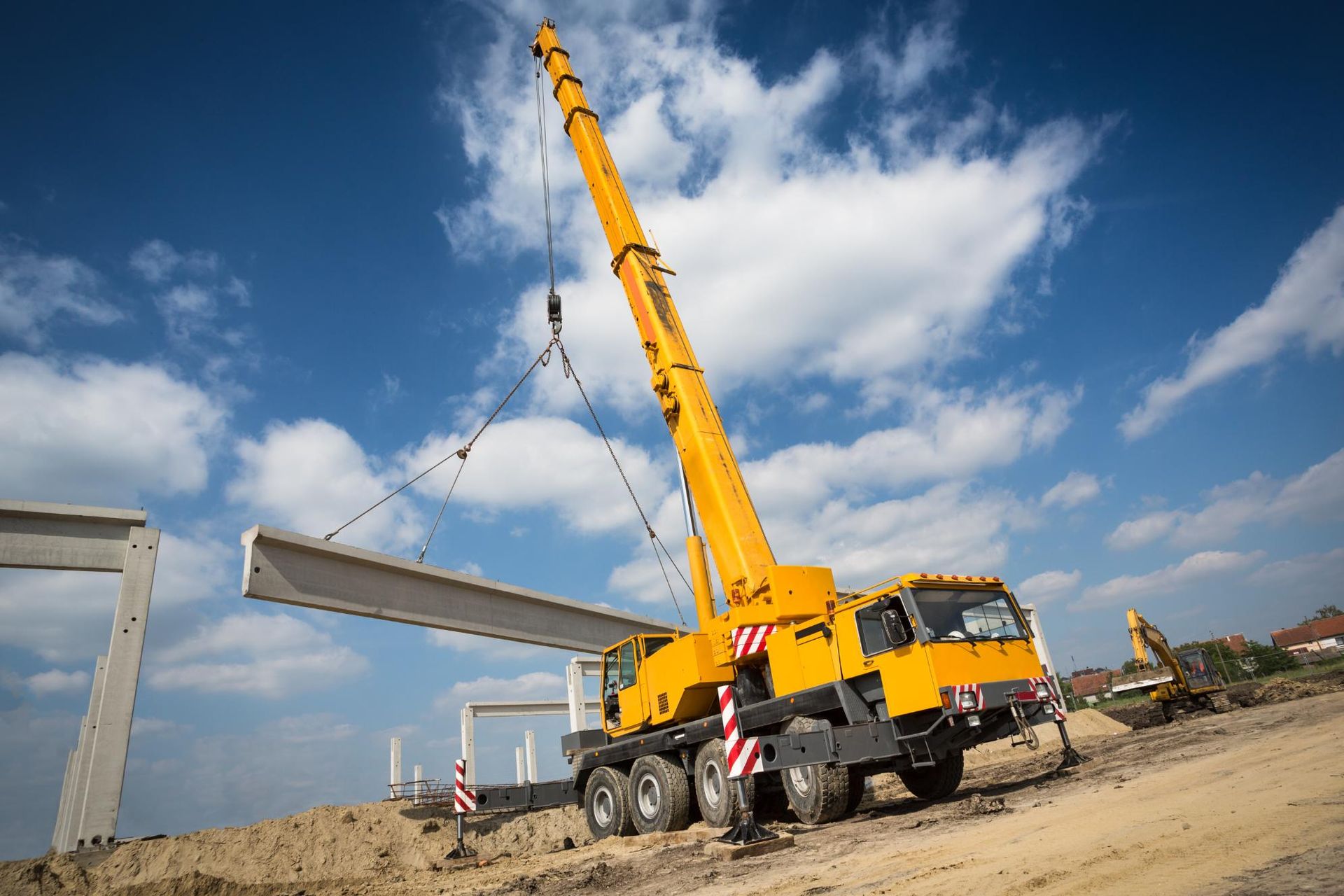 Yellow crane lifting a concrete beam at a construction site under a blue sky with clouds.