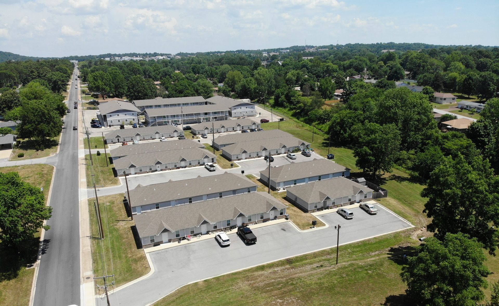 Aerial view of a gray-roofed apartment complex surrounded by trees and a road.