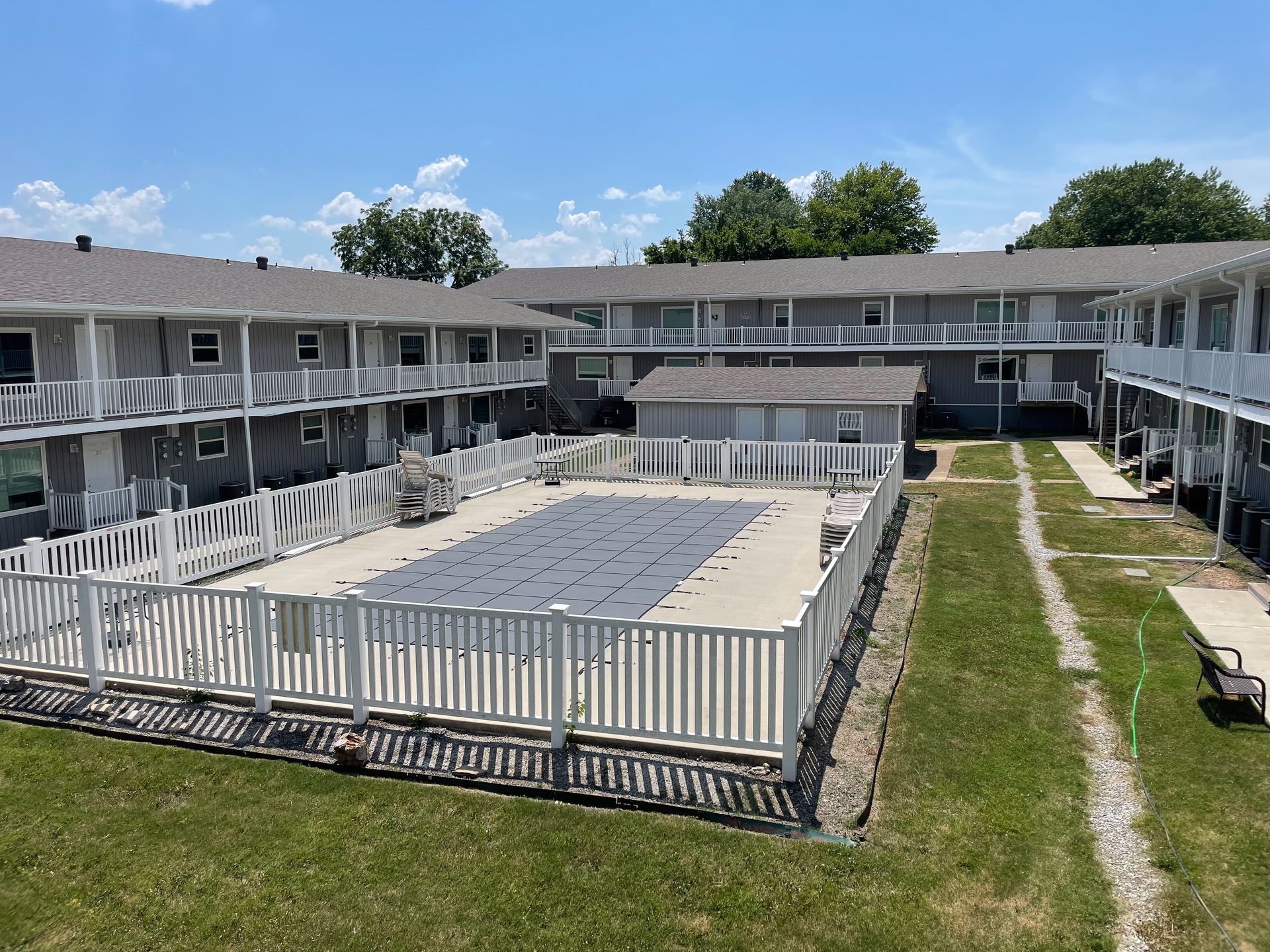 Courtyard of a motel with two-story balconies, white railings, grass, and a fenced pool area under a blue sky