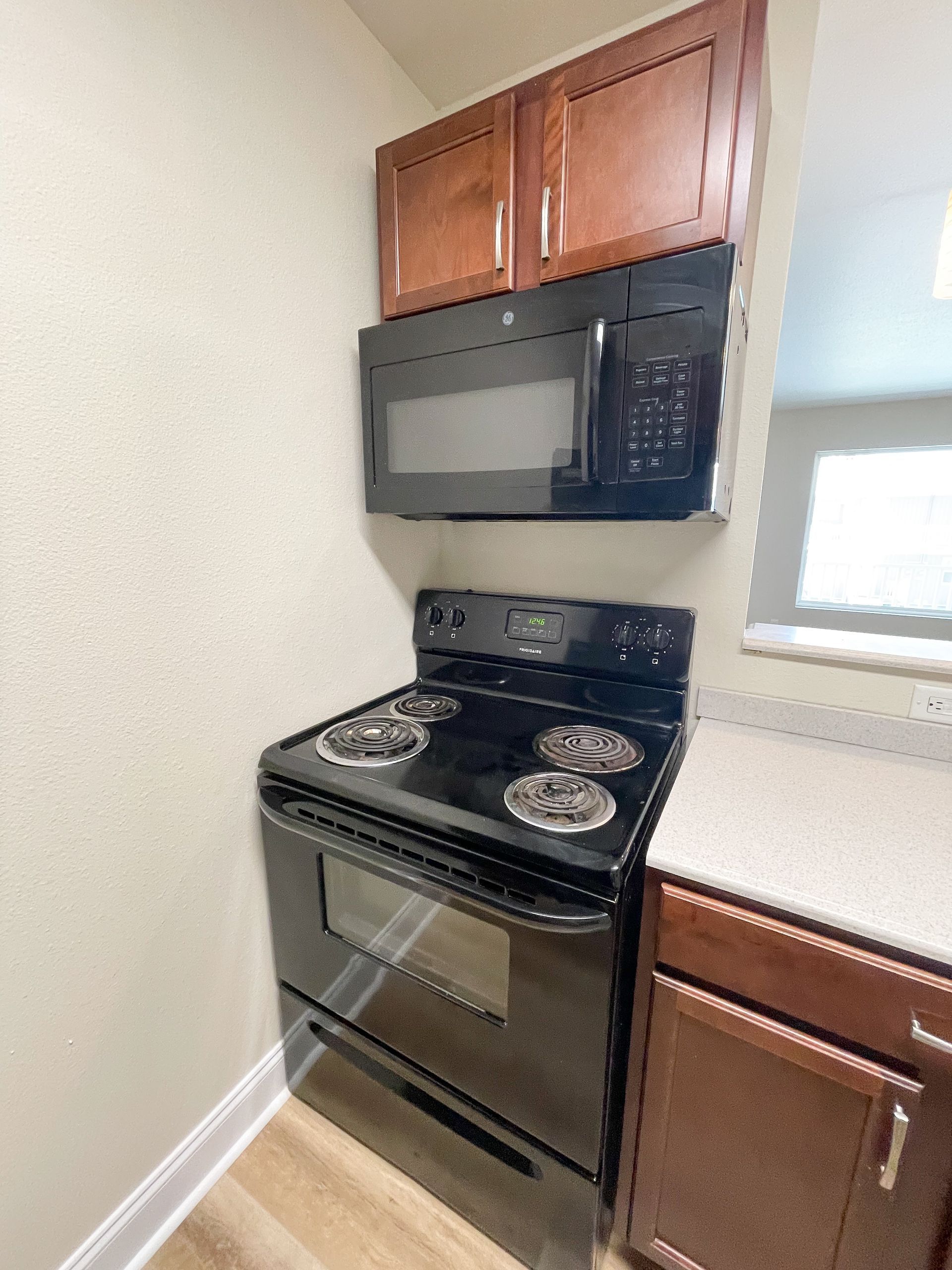 Black kitchen stove and microwave in a small corner with wood cabinets and a window.