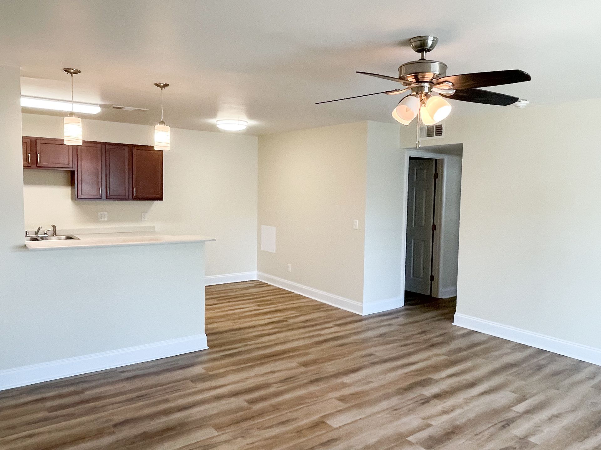Empty open-plan living room with wood-look floors, ceiling fan, and kitchen bar area