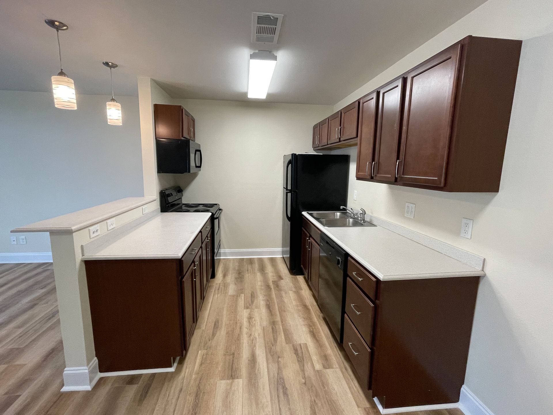 Empty modern kitchen with wood cabinets, white countertops, and a black refrigerator under fluorescent lighting