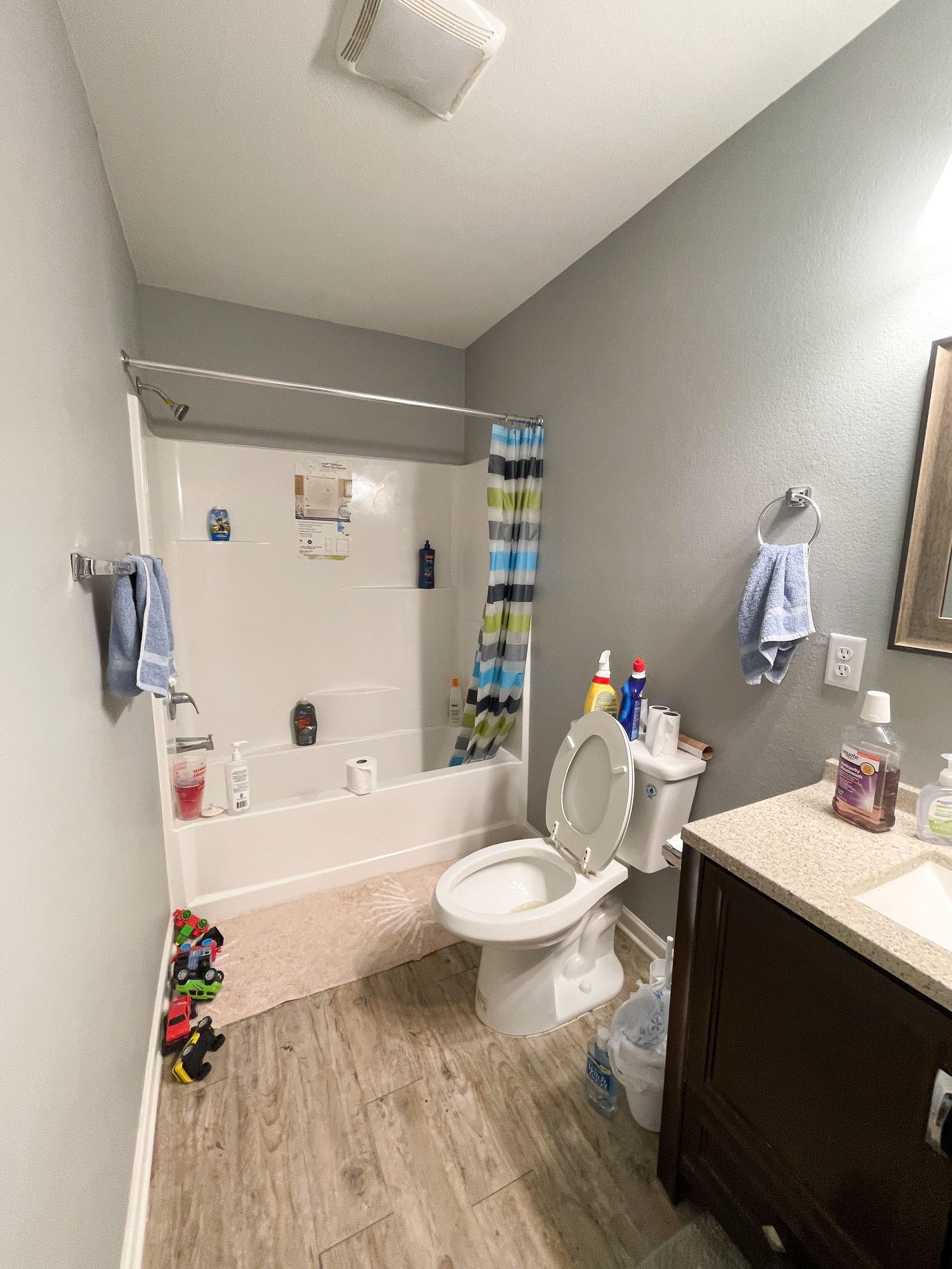 Small bathroom with a tub, toilet, vanity, and towels on gray walls and wood-look floor.