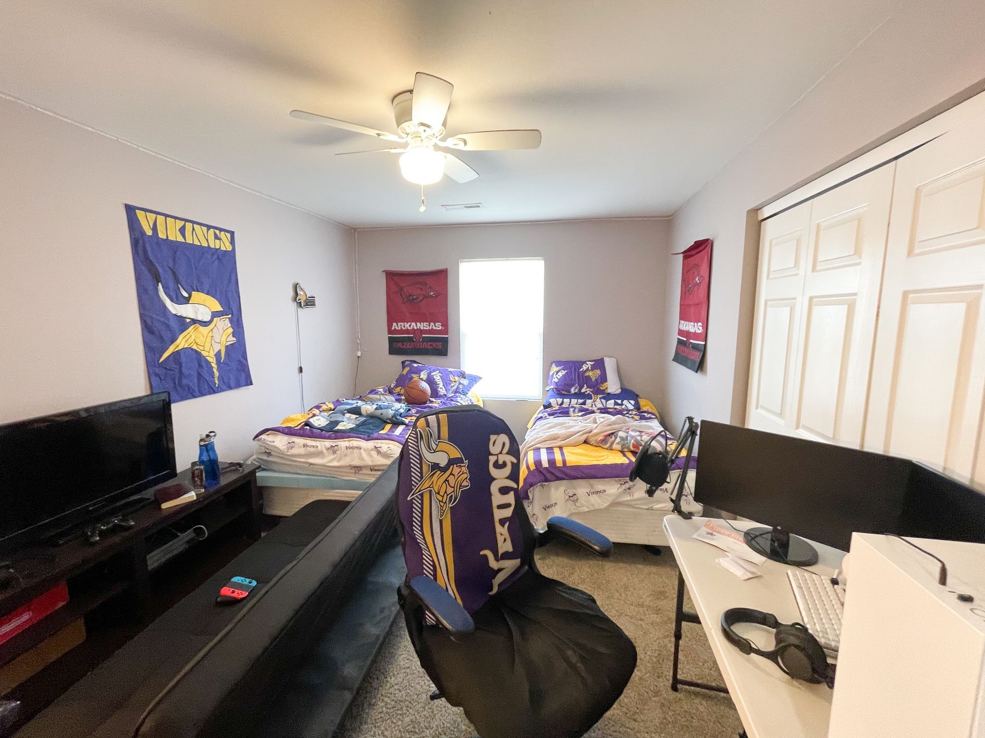 Small bedroom with two twin beds, black desk and monitor, fan ceiling light, and sports wall decor