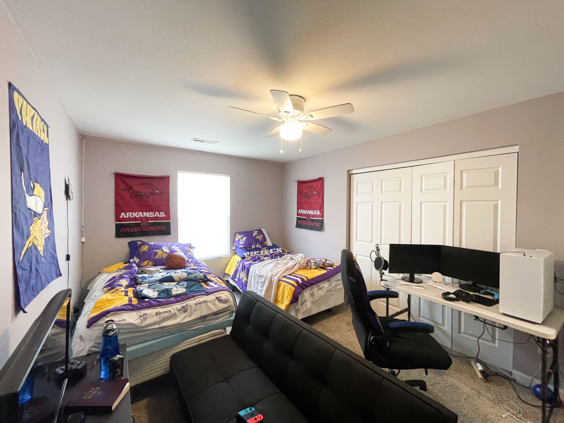 Bedroom with two beds, black couch, desk with computer, and ceiling fan in a bright room.