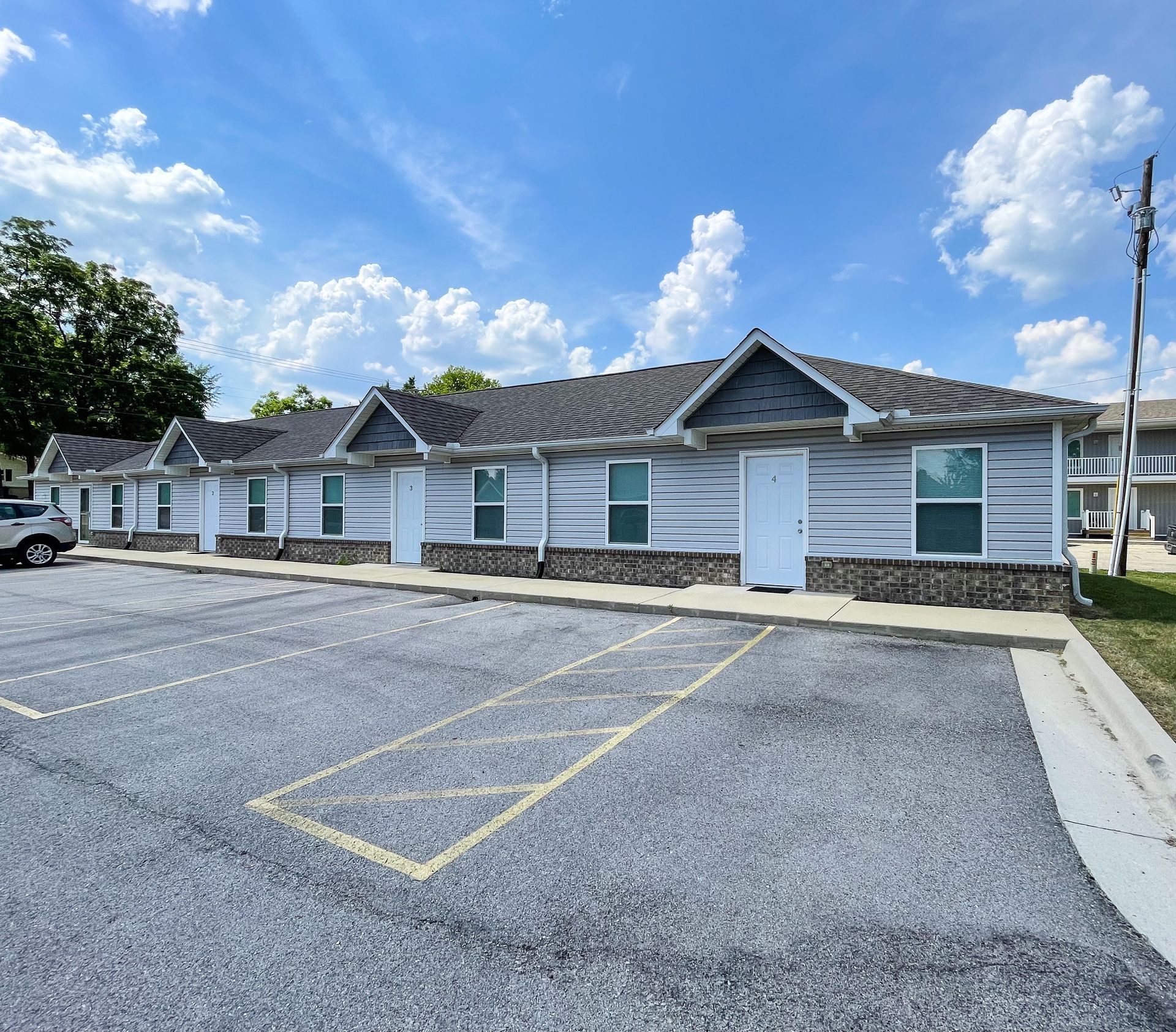 Low, single-story brick building with white siding, blue shutters, and parking spaces in front under a blue sky