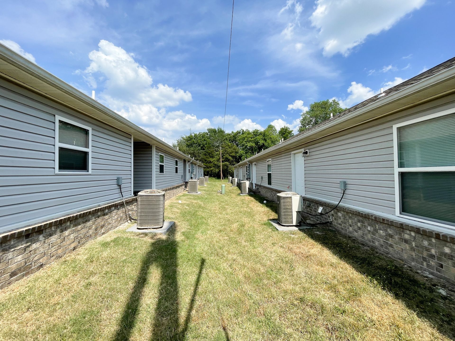 Narrow grassy alley between two white-sided houses under a blue sky, with air conditioners along both walls.