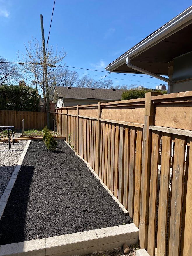 Wooden fence alongside a black mulch garden bed next to a building; bright blue sky.
