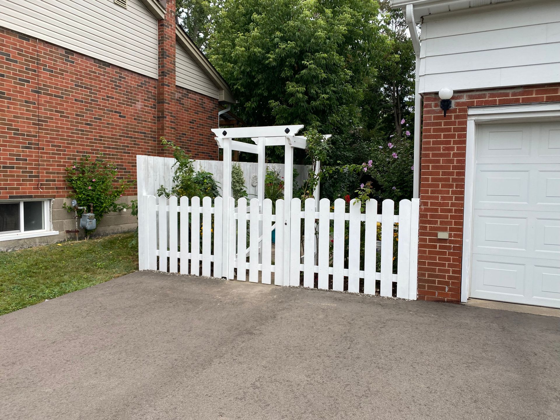 White picket fence with gate in front of a garden and white pergola. Brick building to the left and garage to the right.