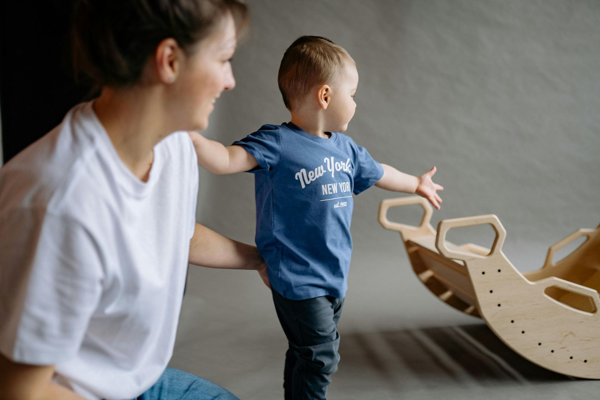 Woman smiles, supports a toddler reaching towards a wooden rocker in a gray-walled room.