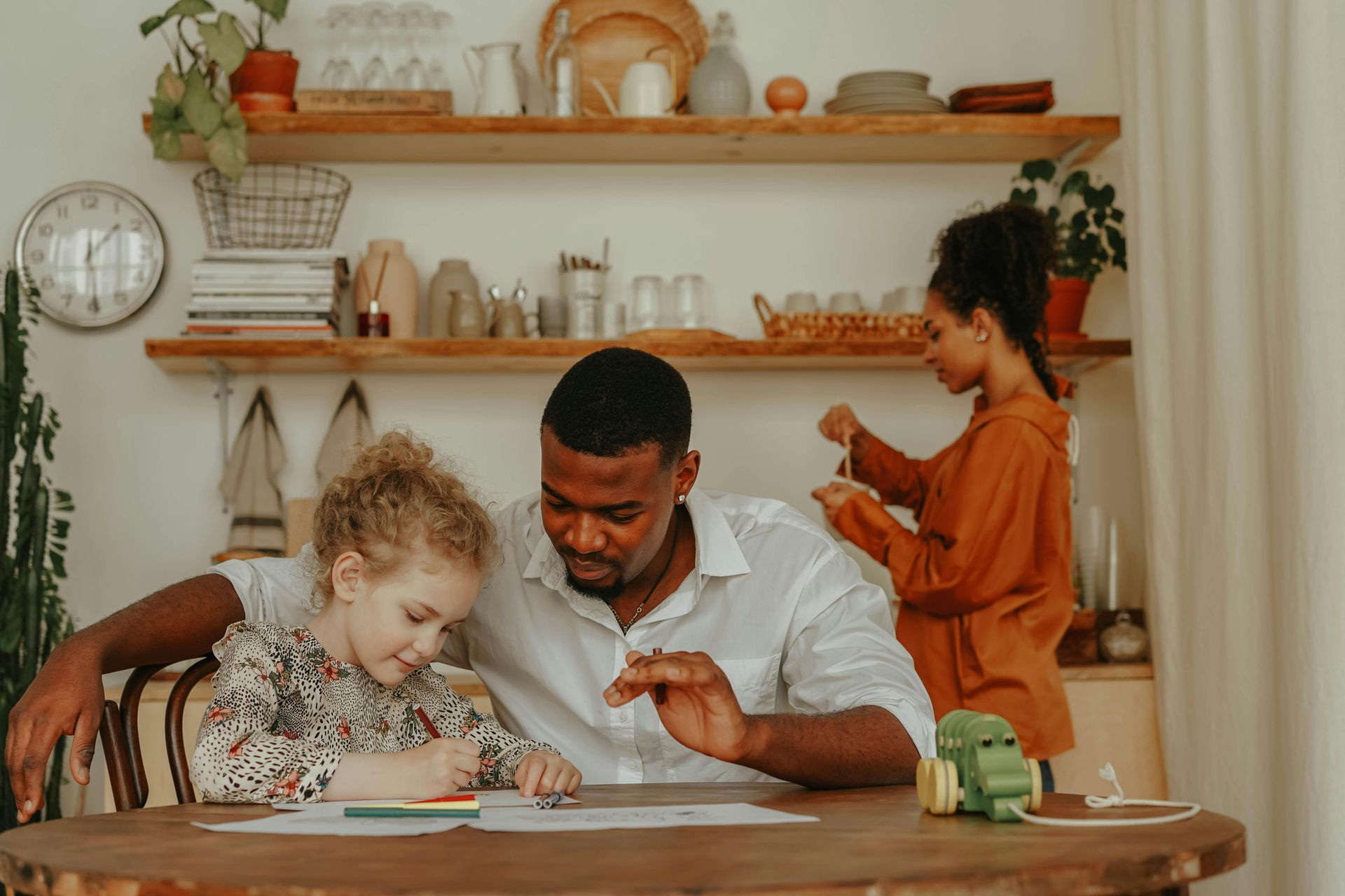 Father and child drawing together at a wooden table, woman in the background near a shelf.
