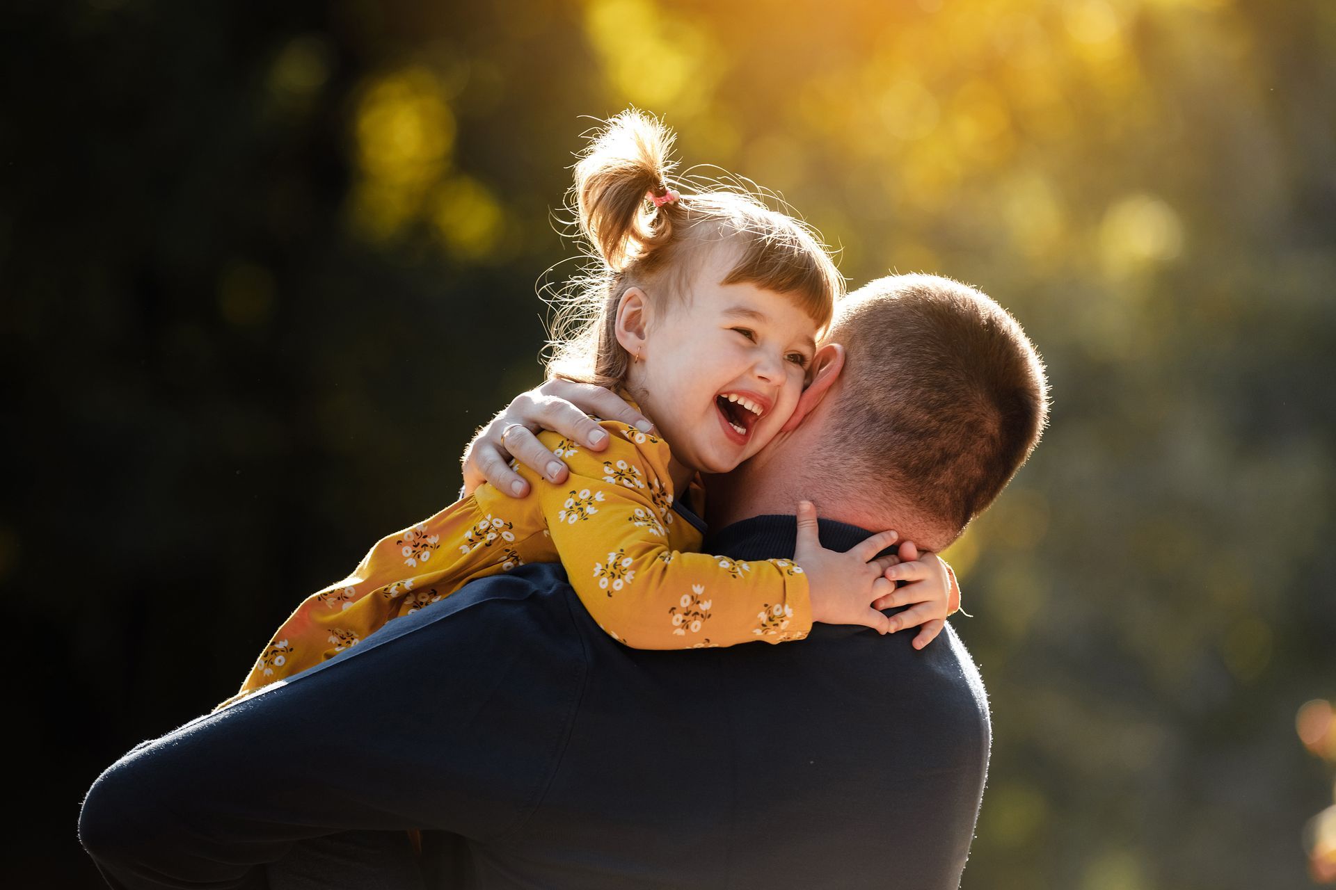 Man holding laughing child, outdoors; golden sunlight.