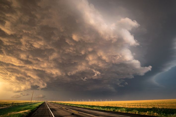 A dark, dramatic storm cloud looms over a rural road and golden wheat fields at sunset.