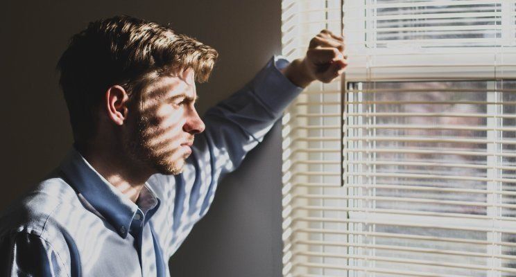 A man is looking out of a window with blinds.