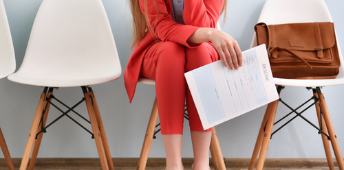 Woman in red suit holding a document, sitting in a chair, waiting for interview. Bag on the chair next to her.