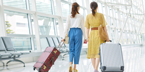 Two women, luggage in tow, walk through an airport terminal toward a bright window.