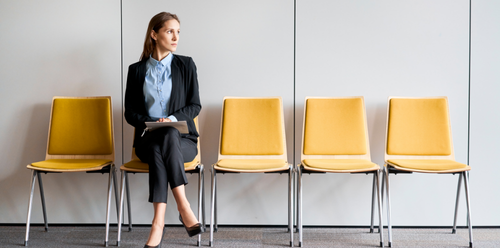 Woman in business attire sits in a yellow chair, looking thoughtful, waiting in an office setting.