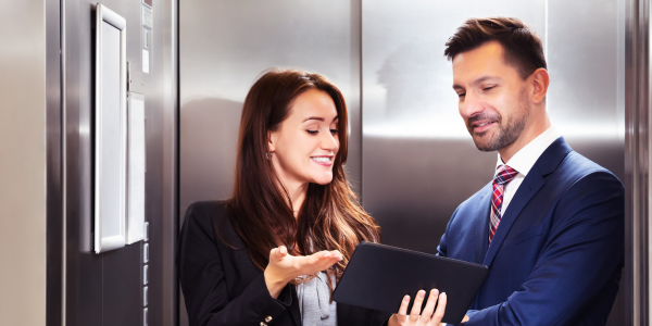 A man and a woman are standing in an elevator looking at a laptop.