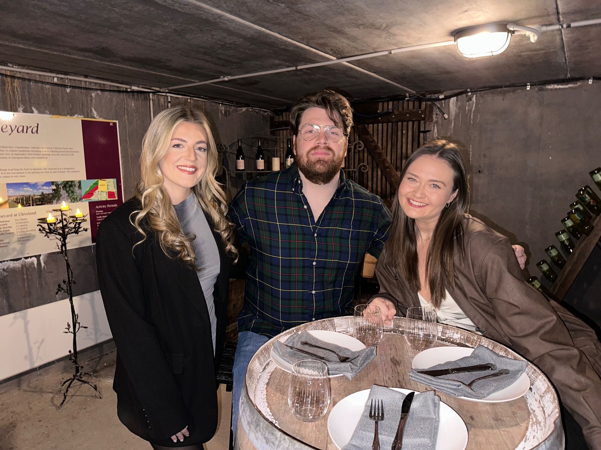 A man and two women are posing for a picture in a wine cellar.