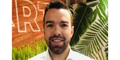 A man with a beard is smiling in front of a wall with palm leaves.
