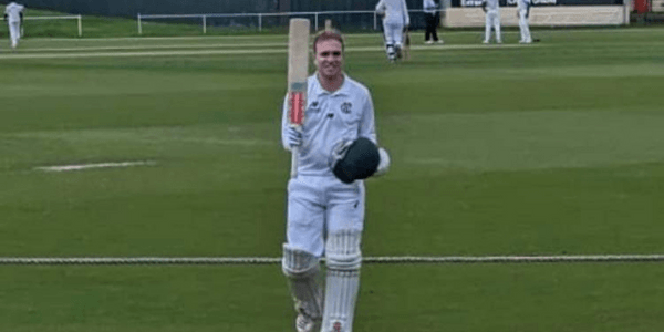 A man is walking on a cricket field holding a bat and helmet.