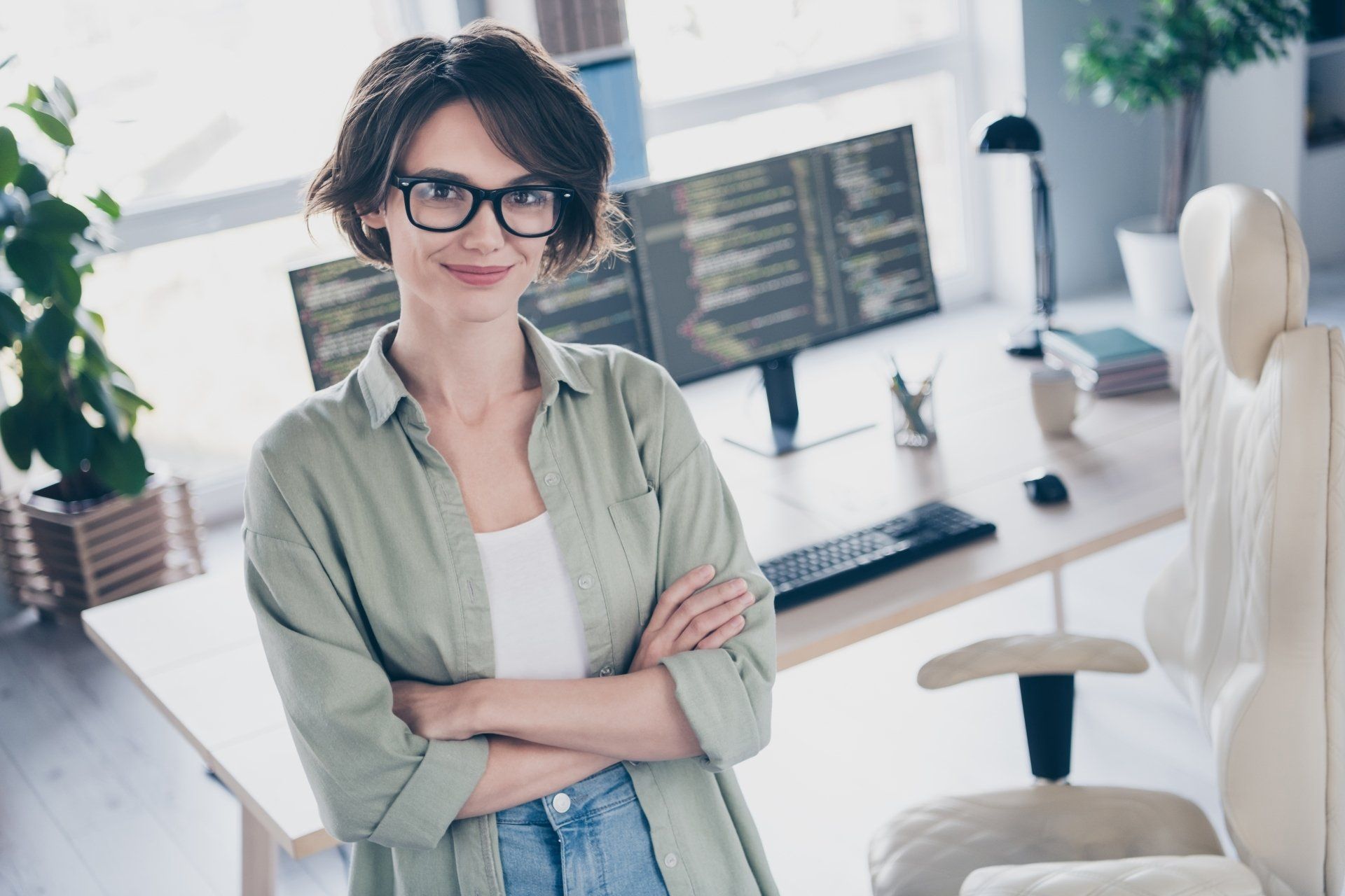 A woman wearing glasses is standing in front of a computer desk with her arms crossed.