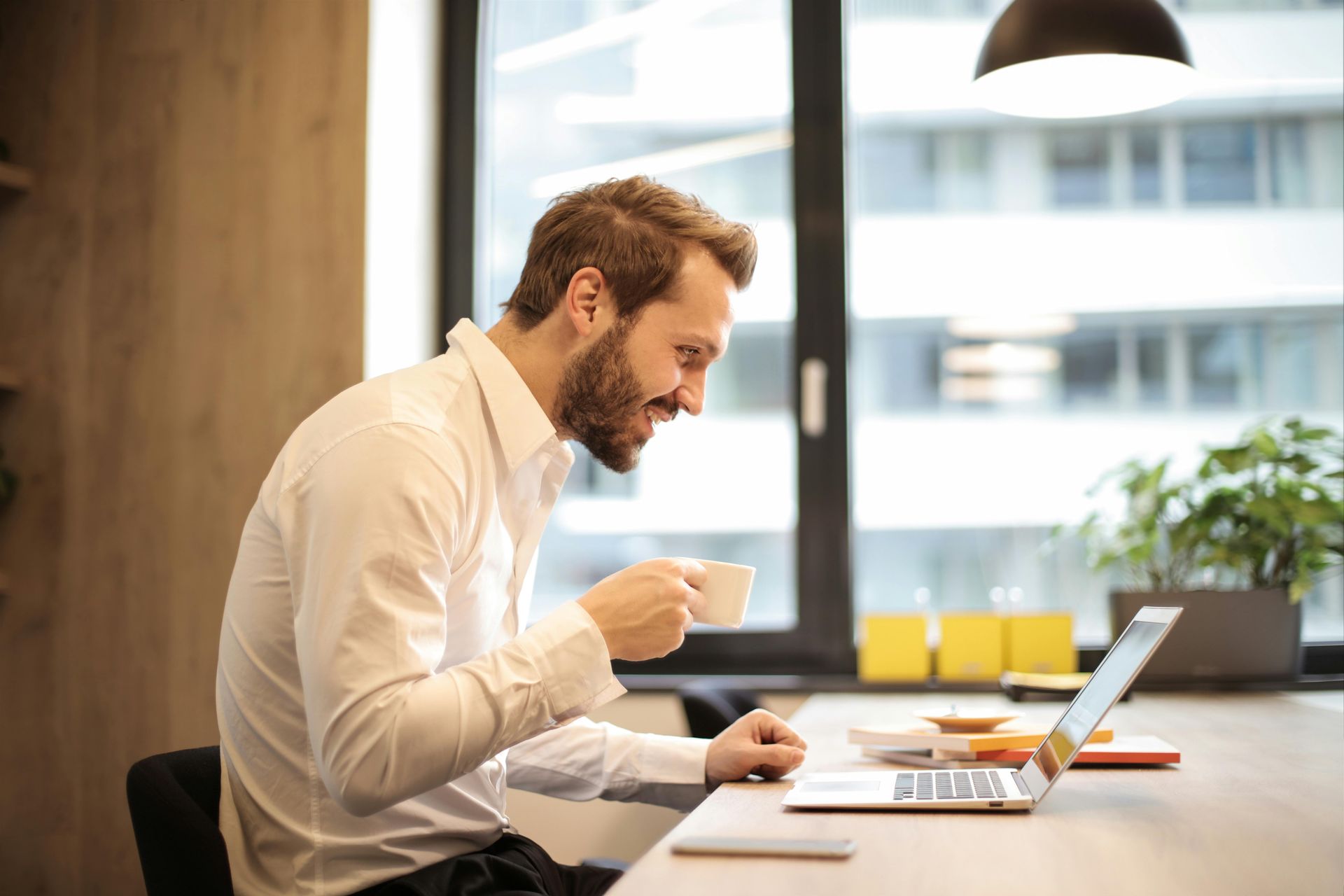 A man is sitting at a table drinking coffee and using a laptop computer.