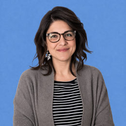 Persona sorridente con occhiali e capelli scuri su sfondo blu, che indossa una camicia a righe e un cardigan grigio.