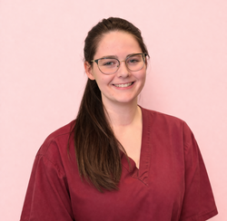 Persona sorridente con occhiali e lunghi capelli scuri, che indossa una camicia bordeaux su sfondo rosa.