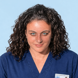 Primo piano di una persona in camice blu con capelli scuri e ricci, su sfondo azzurro.
