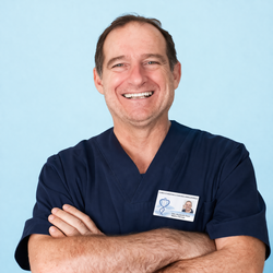 Operatore sanitario sorridente in camice blu scuro, con le braccia incrociate e un cartellino identificativo su sfondo azzurro.