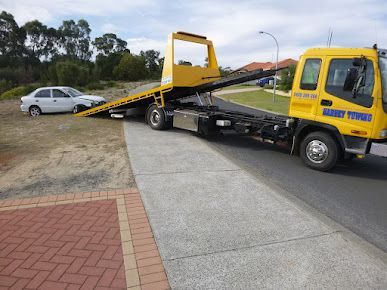 Yellow tow truck loading a white car on a ramp beside a curb on a roadside.