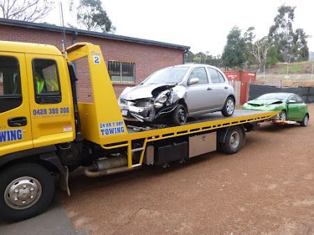 Tow truck hauling a damaged silver car on a flatbed, with a green car behind it on a dirt road.