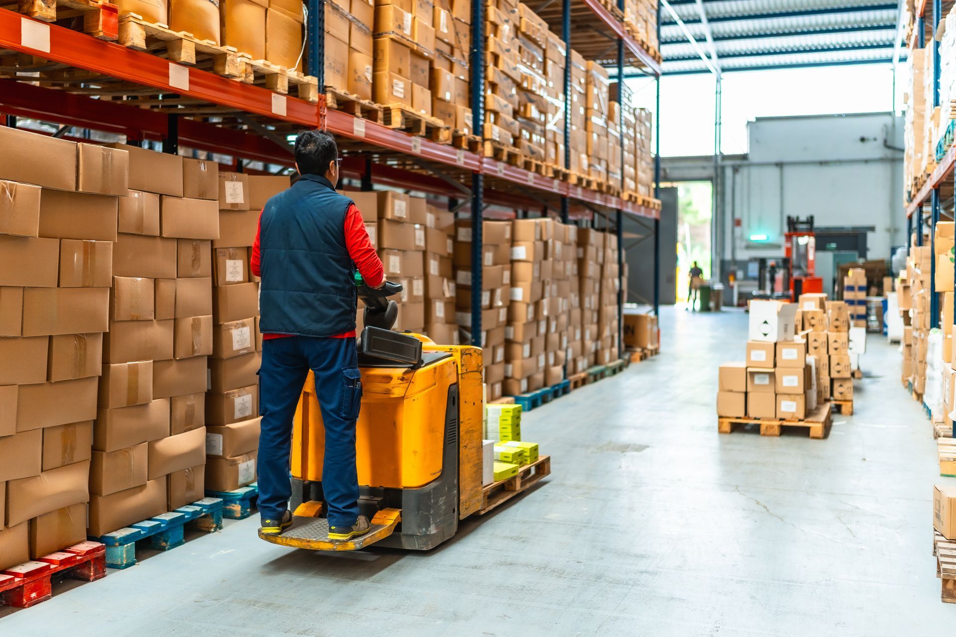 Warehouse worker operating a yellow pallet jack beside stacked cardboard boxes in a storage aisle