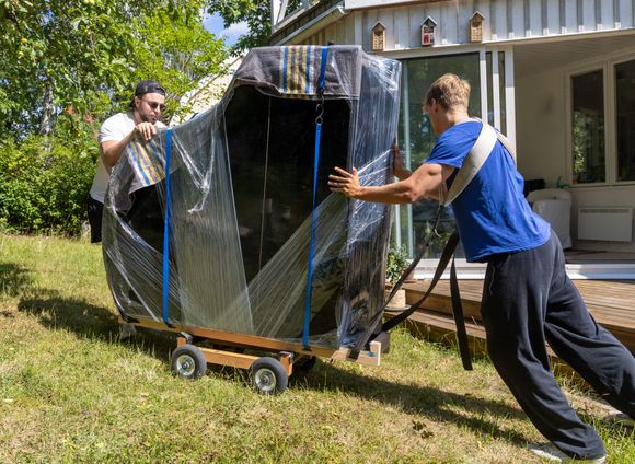 Two people pushing a large wrapped object on a dolly across a yard