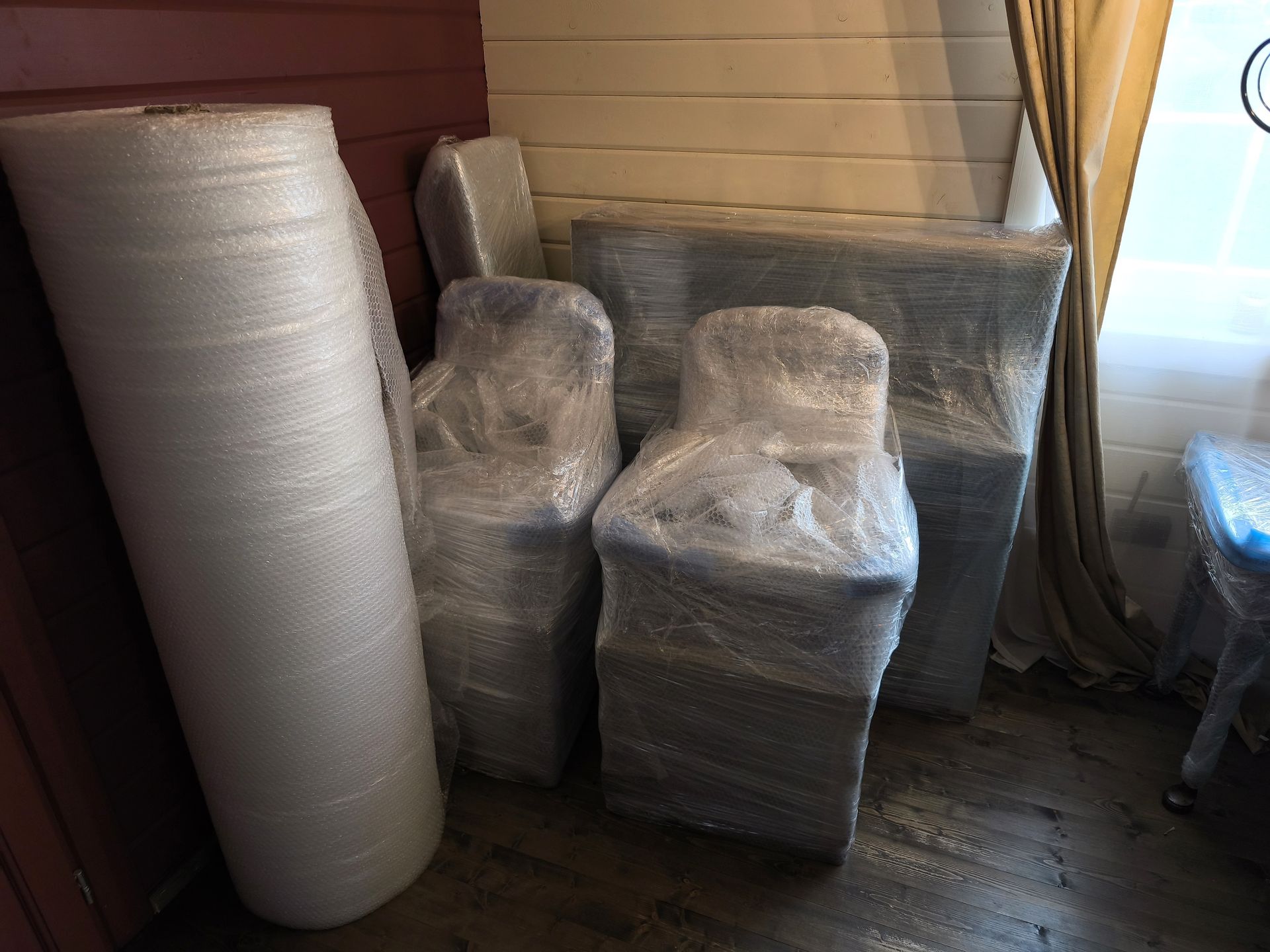 Wrapped furniture and a tall roll of bubble wrap stored in a room by a window and curtains