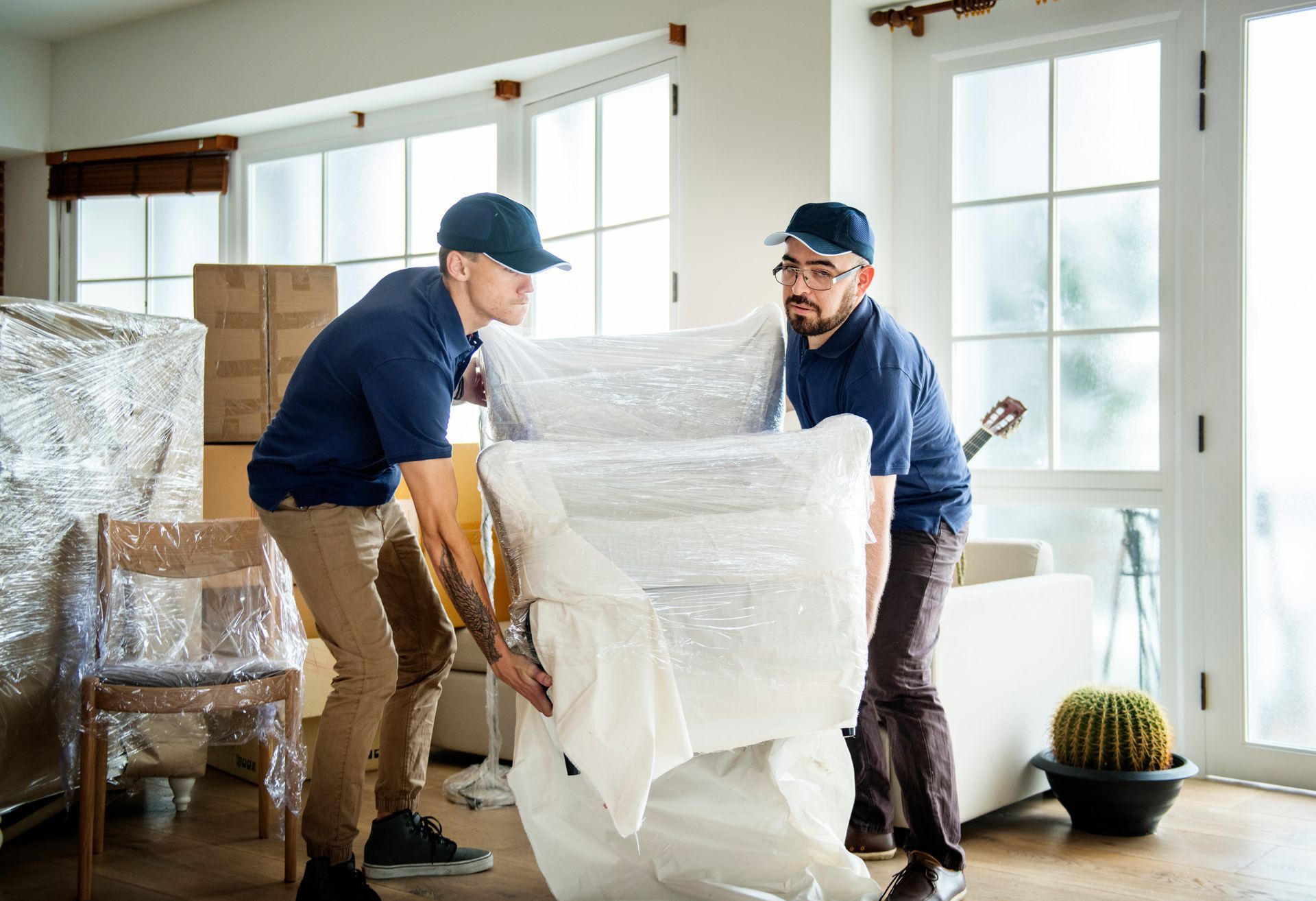 Two movers carrying a wrapped sofa through a bright living room with stacked boxes and large windows