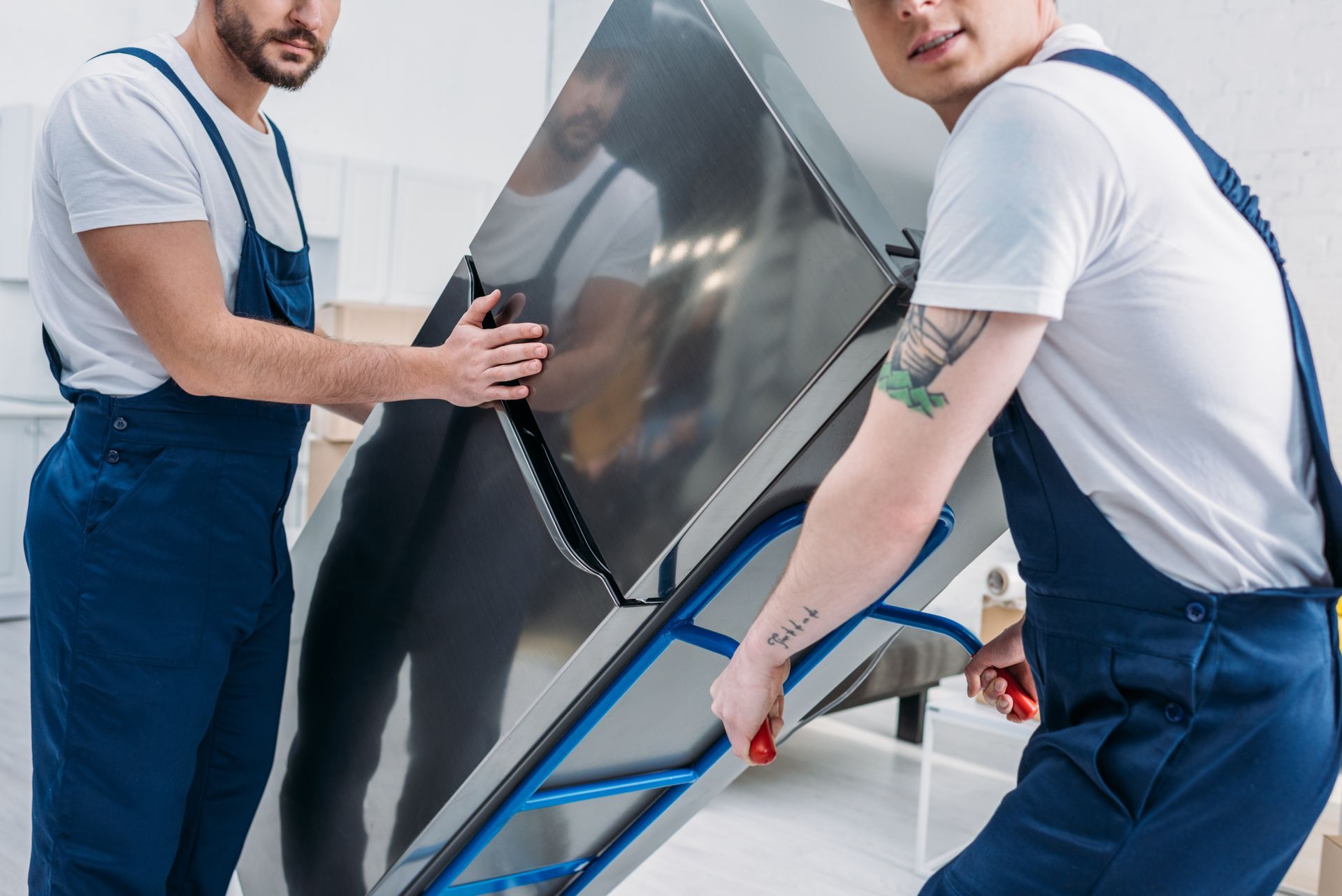 Two movers in blue overalls carrying a large stainless steel appliance on a dolly indoors