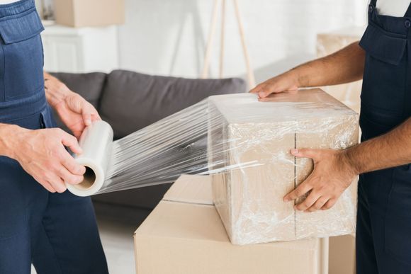 Two people wrapping a cardboard box with clear plastic film in a moving setting
