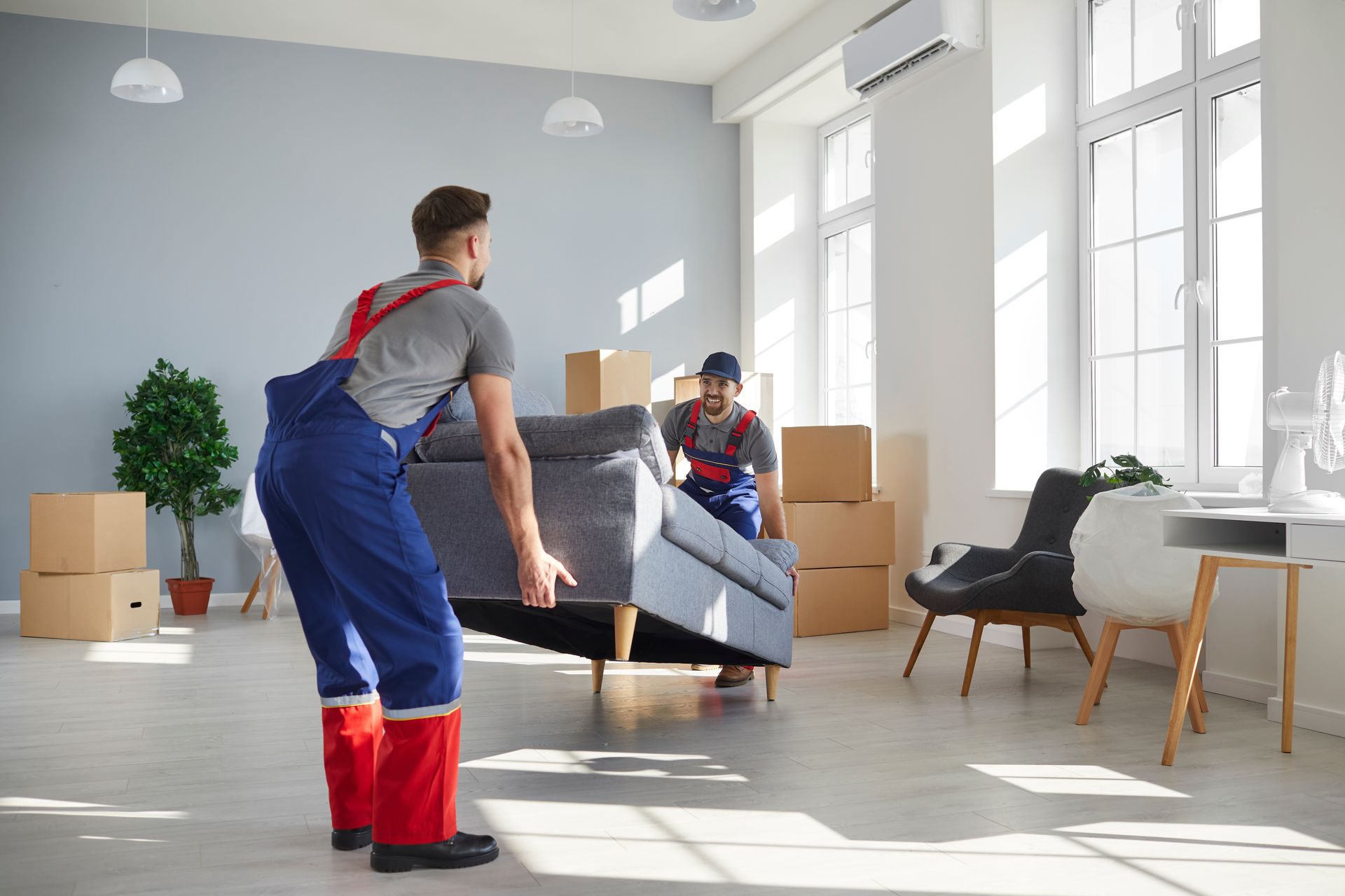 Two movers carrying a gray sofa in a bright living room with boxes and chairs nearby.