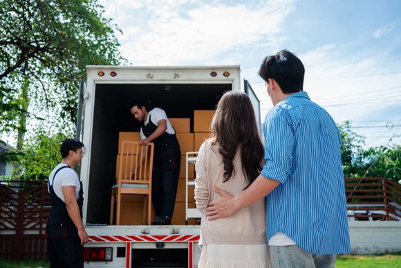 People unloading furniture from a moving truck outdoors, with boxes and chairs inside.